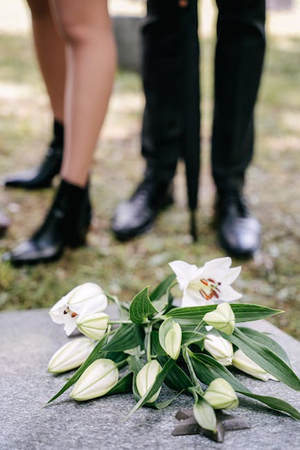 A delicate bouquet of white lilies with open blossoms revealing their orange stamens, surrounded by closed buds and lush green leaves, resting on a stone surface outdoors. In the background, two indiv