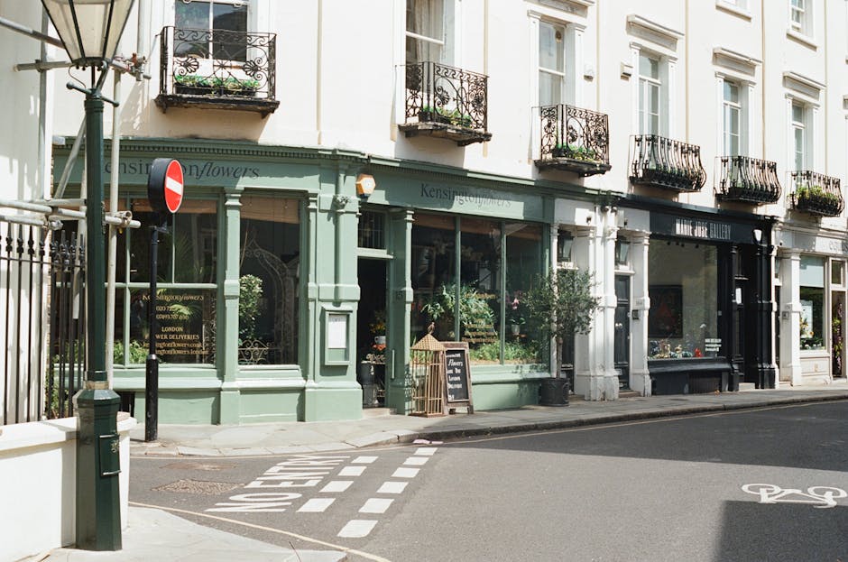 A floral shop with a pale green façade and large display windows showcasing fresh flowers, situated on a street corner with a black wrought iron balcony above. The shop has a small chalkboard sign ou