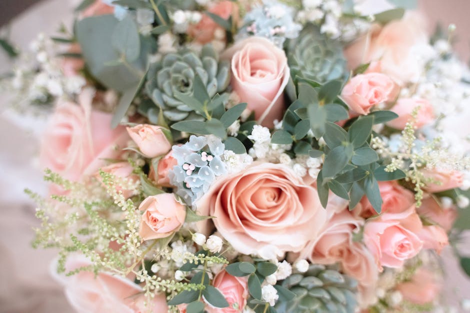 A close-up view of a floral bouquet featuring large, blush pink roses with delicate, layered petals, complemented by smaller pink spray roses. The arrangement includes silvery-green eucalyptus leaves,