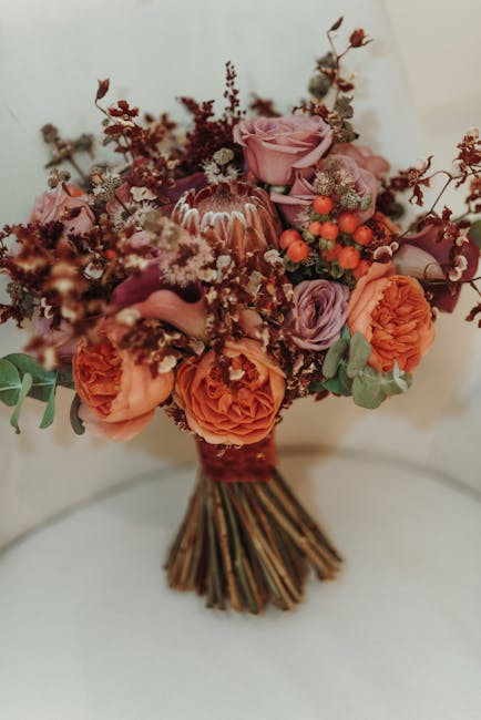 A bouquet arrangement featuring soft pink roses, vibrant orange ranunculus, and small clusters of red berries, complemented by sprigs of dusty miller and eucalyptus leaves. The flowers are freshly cut