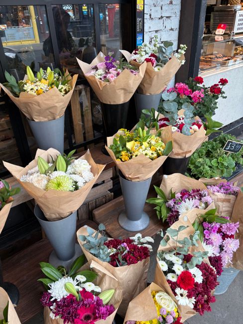 A display of various floral arrangements outside a shop, featuring bouquets of fresh flowers wrapped in brown kraft paper and placed in tall, elegant grey vases. The flowers include white lilies, yell