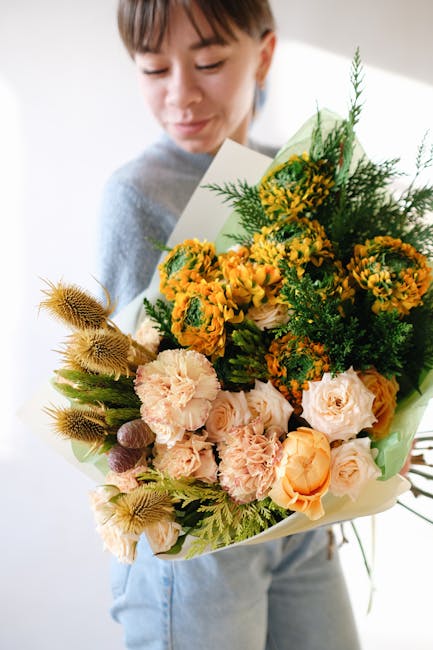 A young woman holding a large bouquet of fresh flowers wrapped in light green paper, featuring a mix of peach, yellow, and cream blooms including roses, carnations, and small yellow daisies, accented 